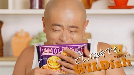 坊主頭の男性が、紫色のパッケージに入った「WILDish」という食品を手に持ち、目を閉じて匂いを嗅いでいるような表情をしている。キッチンらしき場所で撮影された写真。